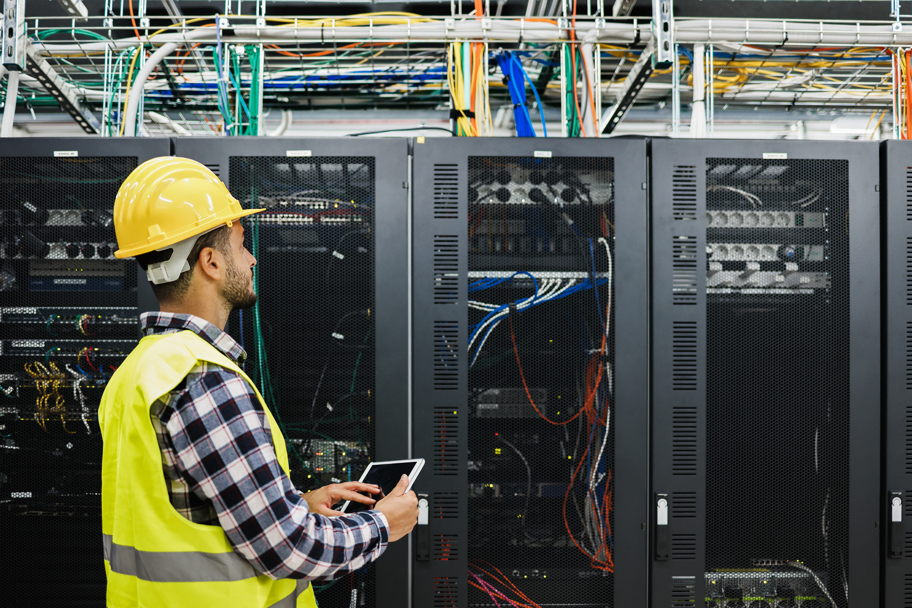 A male technician wearing a yellow hard hat, safety vest, and plaid shirt stands in a server room holding a tablet. He faces a row of large black server cabinets filled with cables and electronic equipment, with colorful wiring visible overhead.