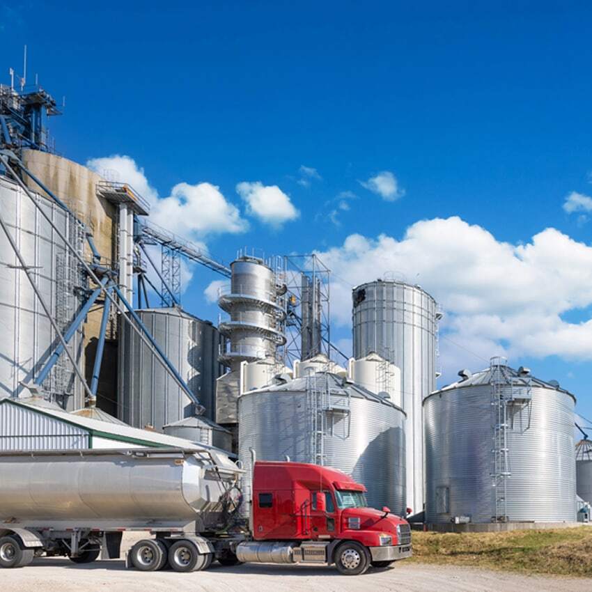 A red semi-truck with a silver tanker trailer is parked in front of large silver grain silos and industrial structures under a blue sky with scattered white clouds.