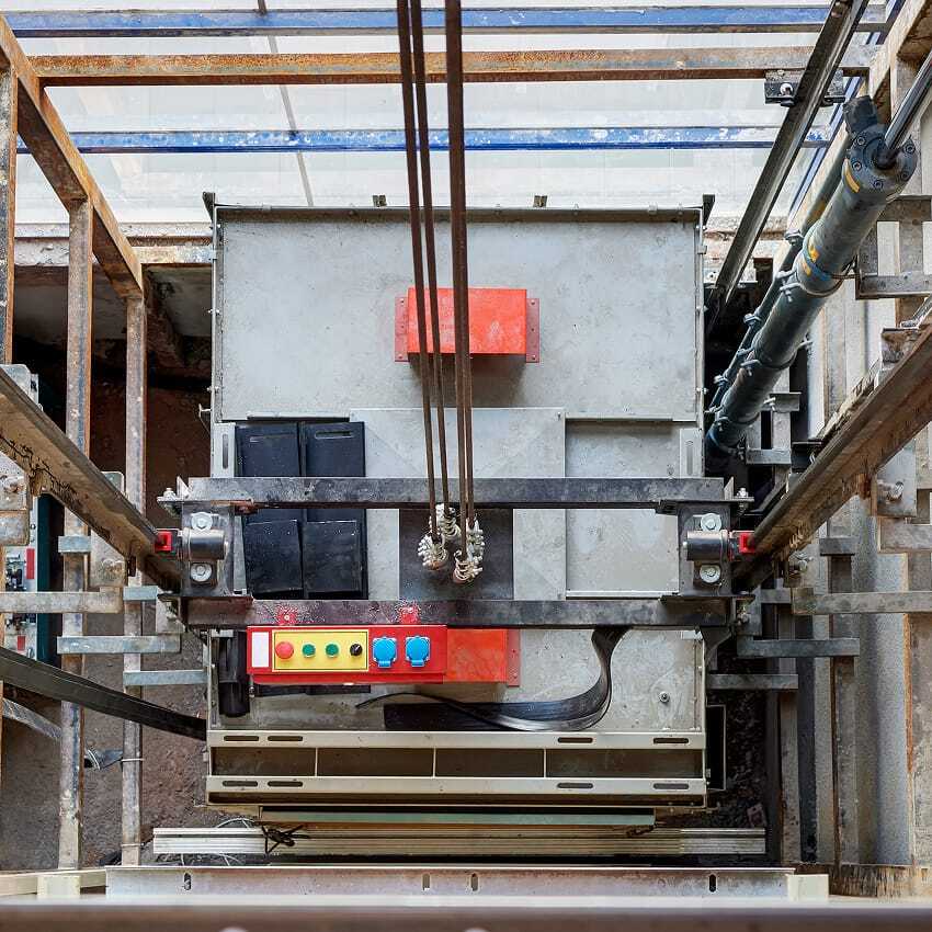 View from above of an elevator shaft with visible rails, pulleys, and cables connected to the top of the elevator car. The car has a red control box with colored buttons on top. The elevator is under maintenance.