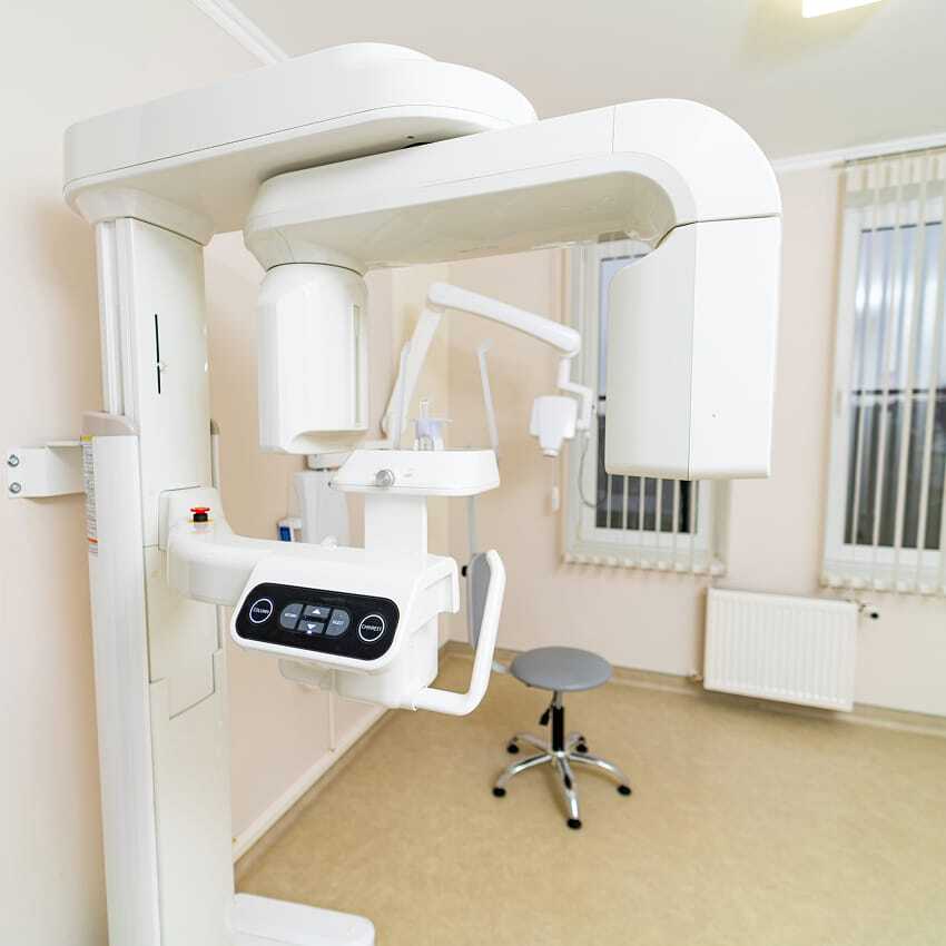 A panoramic dental X-ray machine in a bright, clean dental office with beige floors, white walls, vertical blinds on the windows, and a gray stool.