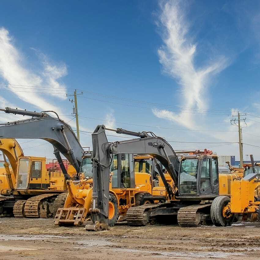 A row of construction vehicles, including excavators, bulldozers, and a road roller, parked on a dirt lot under a blue sky with wispy clouds. Buildings and utility poles are visible in the background.