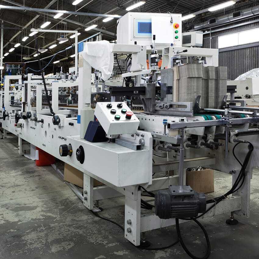 A large industrial packaging machine with control panels and conveyor belts stands in a factory setting, surrounded by various equipment and stacks of cardboard. Overhead lights illuminate the spacious, organized workspace.