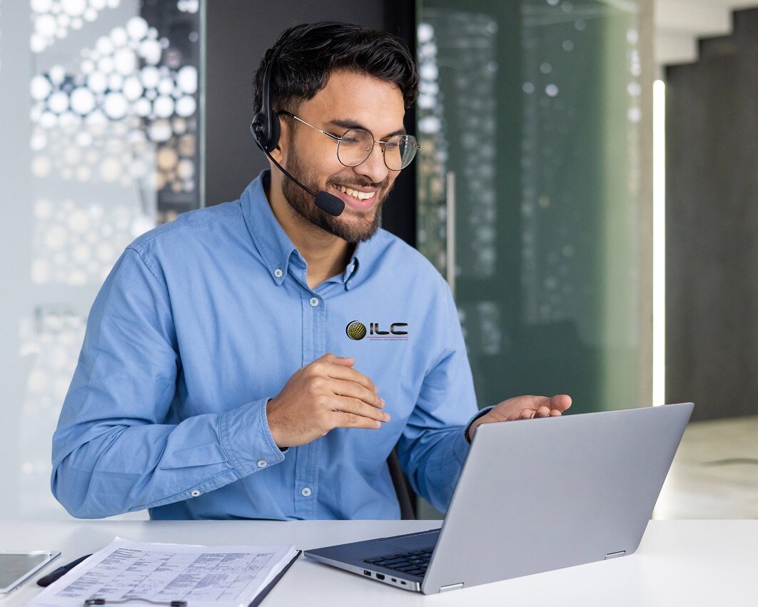 A smiling man in a blue shirt and glasses sits at a desk using a laptop, wearing a headset. A clipboard rests nearby. The setting is bright and modern.