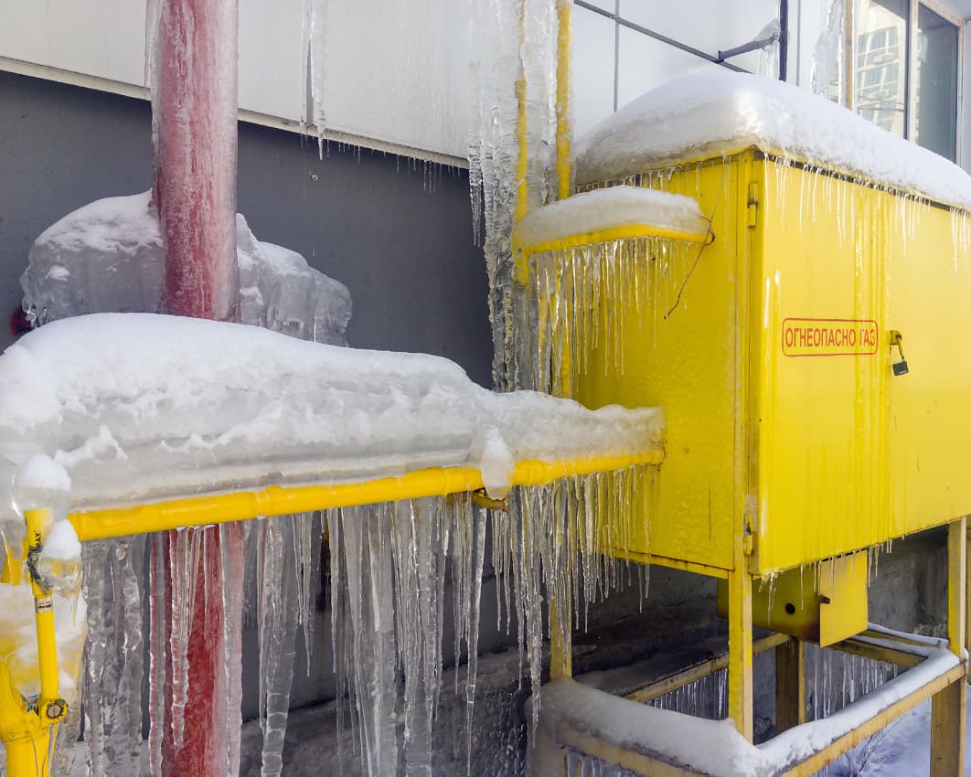A yellow gas control box and pipes, labeled in Russian, are covered in ice and icicles, with snow piled on top. A red vertical pipe and a gray wall are in the background, also coated with ice.