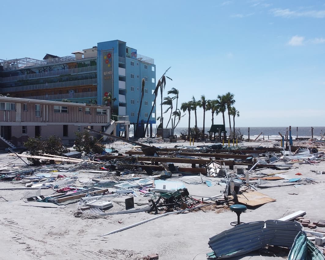 A beachfront area with debris and damaged structures scattered across the sand, palm trees bent or stripped, and a building with visible roof damage in the background under a clear blue sky.