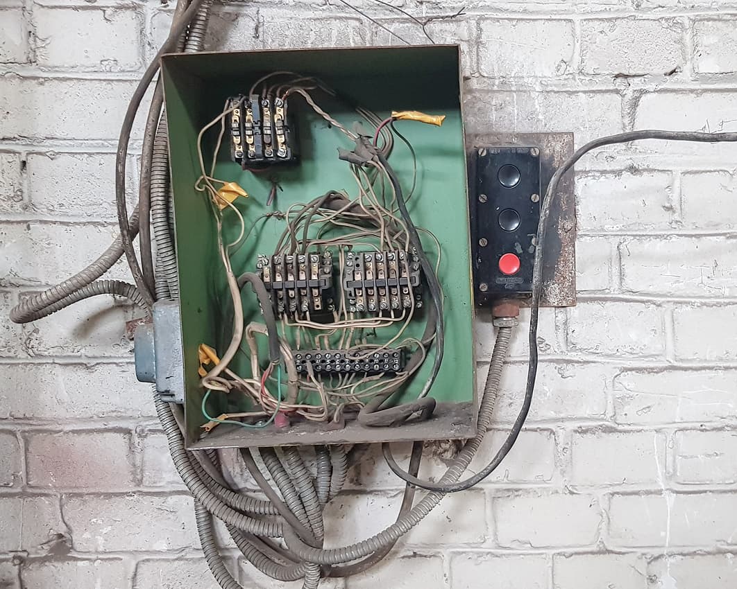 An old electrical panel with tangled, exposed wires and corroded components mounted on a white brick wall. A metal control box with a large red button is beside it. The scene appears neglected and unsafe.