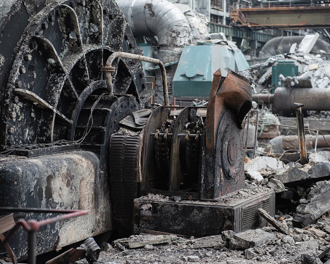 A large, rusted, and partially destroyed industrial machine sits among rubble and debris in a ruined factory or industrial site, with crumbling structures and pipes in the background.