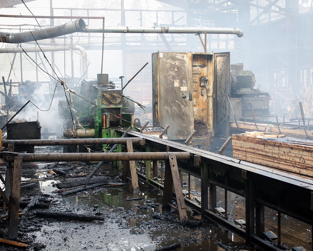 Interior of a damaged industrial factory with burnt machinery, twisted metal, debris, and smoke. The structure appears heavily affected by fire, with charred surfaces and water on the floor.