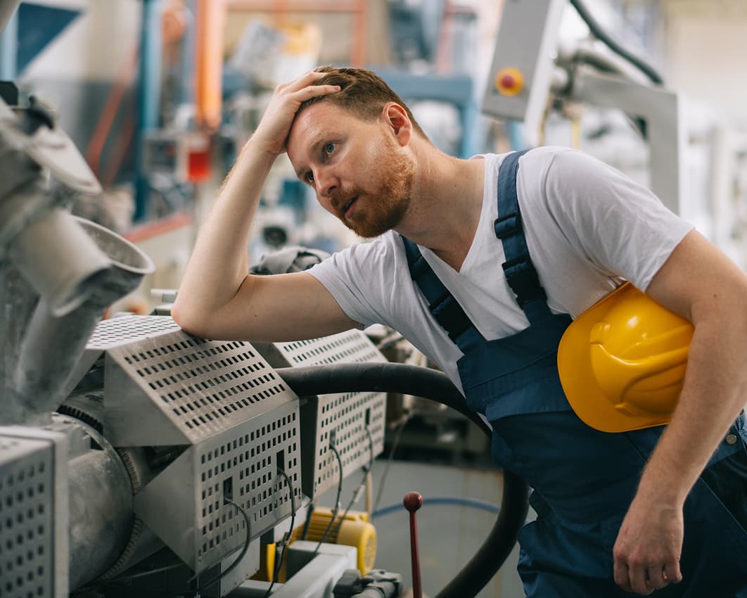 A man in blue overalls and a white t-shirt leans on industrial machinery, holding a yellow hard hat and looking tired or stressed, in a factory setting.