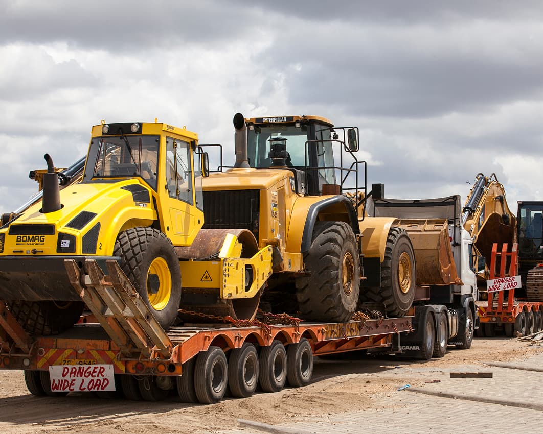Several large construction vehicles, including a yellow road roller, a wheel loader, and an excavator, are loaded onto flatbed trailers on a construction site under a cloudy sky. A sign reads Danger Wide Load.