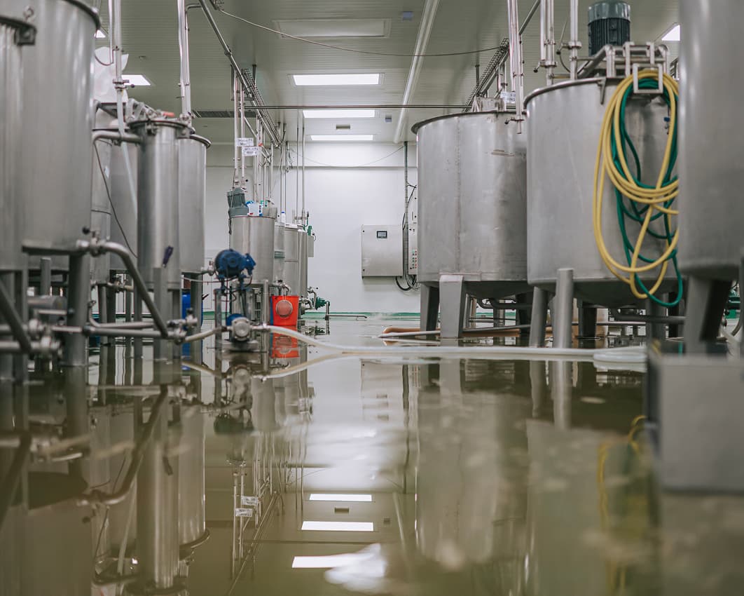Large stainless steel tanks in an industrial facility with water covering the floor, reflecting the equipment and ceiling lights; hoses and pipes are visible throughout the space.