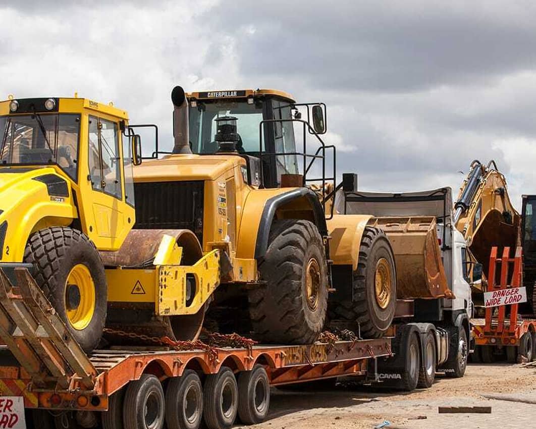 Several large construction vehicles, including a yellow road roller, a wheel loader, and an excavator, are loaded onto flatbed trailers on a construction site under a cloudy sky. A sign reads Danger Wide Load.