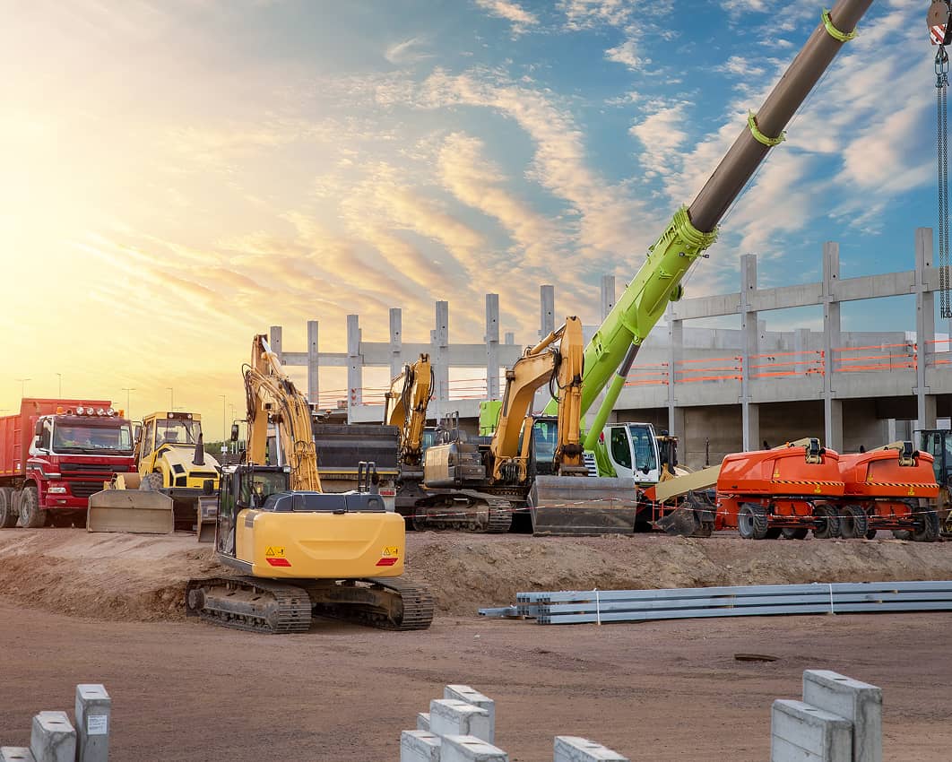 A construction site at sunset with multiple pieces of heavy machinery, including excavators, a crane, a dump truck, and compactors. Concrete columns and a partially built structure stand in the background while machinery is parked on dirt in the foreground under a colorful sky.