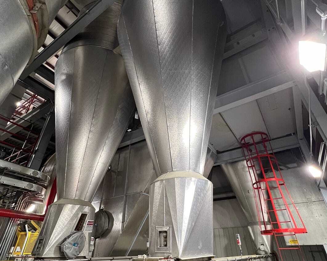 Large industrial metal silos in a warehouse with gray walls and exposed pipes. A red ladder extends vertically. The space appears clean and organized.