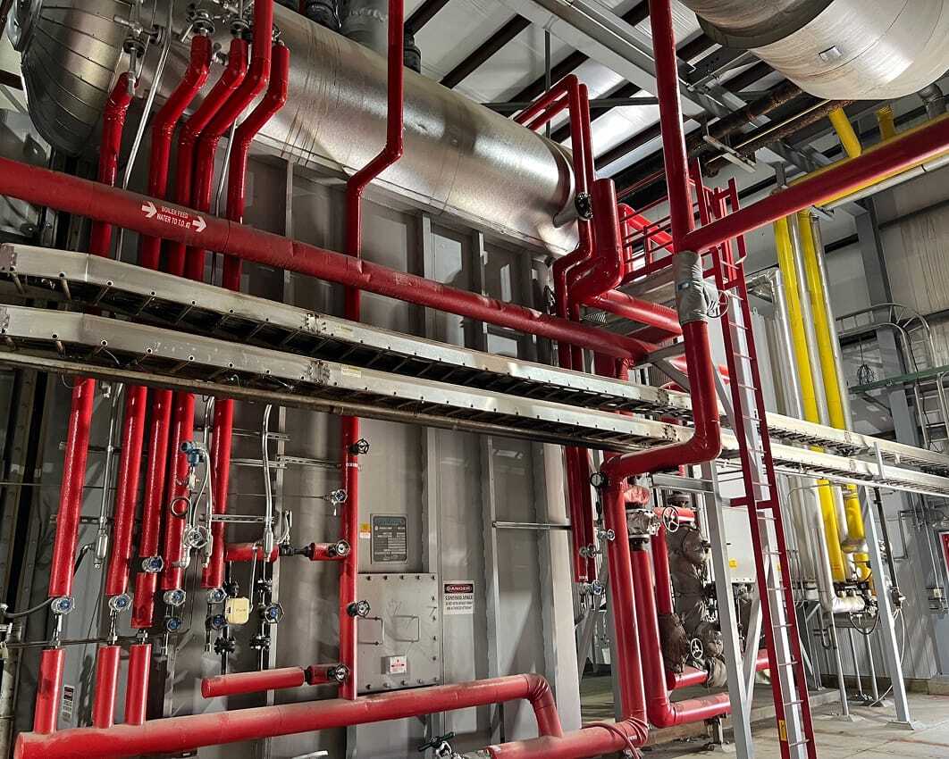 A complex network of industrial pipes in a facility. Red, yellow, and metallic pipes with valves and gauges suggest an active system. Utilitarian tone.