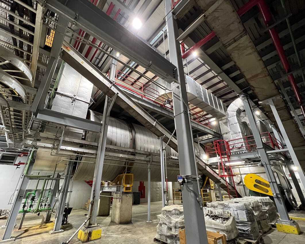 Interior of an industrial facility with large metal ducts, beams, and red railings. The space appears spacious, organized, and illuminated by bright lights.