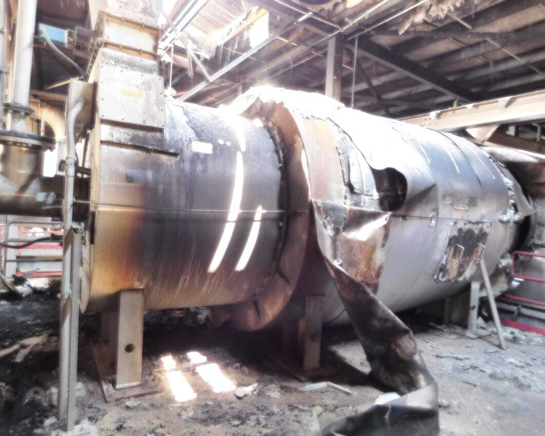 Large, damaged industrial tank inside a warehouse, with scorched metal and debris around, suggesting a fire incident. Sunlight filters through roof gaps.