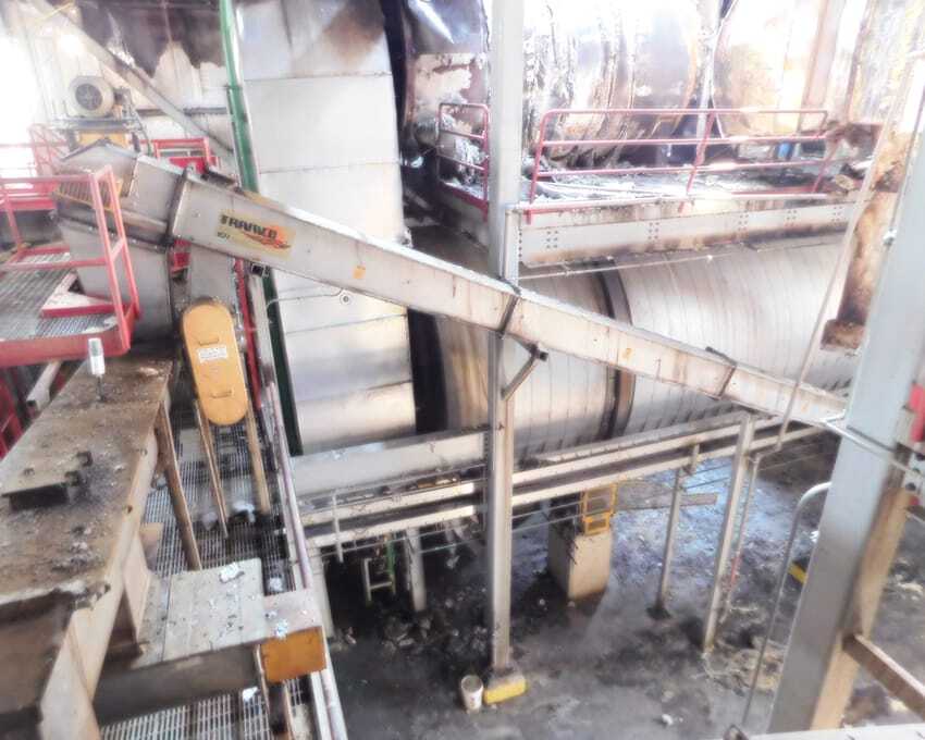 Man in safety gear and a hard hat inspects industrial machinery in a factory. The scene includes red metal structures and a yellow safety railing.