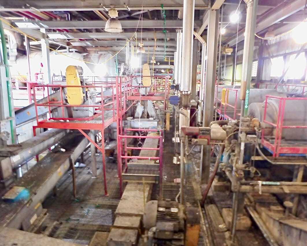 Industrial interior with metal walkways and red railings. Machinery and pipes fill the space, creating a complex, busy atmosphere. Bright lighting.
