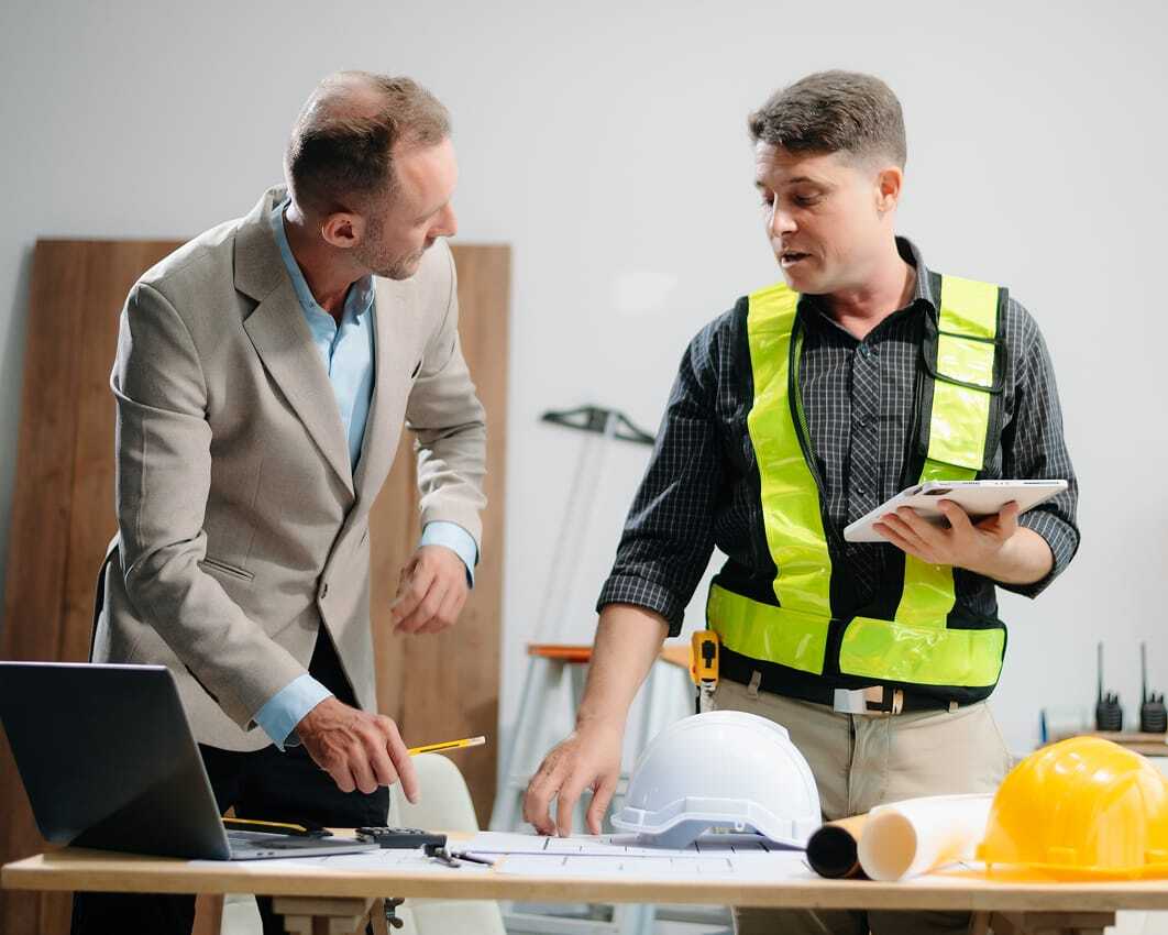 Two men discuss construction plans at a table in an office setting. One man wears a beige suit and gestures toward blueprints on the desk, while the other wears a reflective safety vest and holds a tablet. Hard hats, rolled plans, and a laptop are visible on the table.