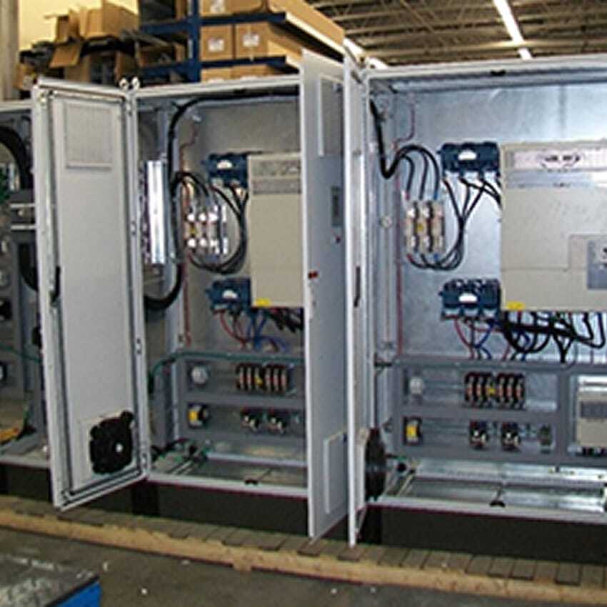 Three large electrical control panels with open doors reveal wiring, circuit breakers, relays, and various electrical components inside. The panels are installed in an industrial setting with shelves and boxes in the background.