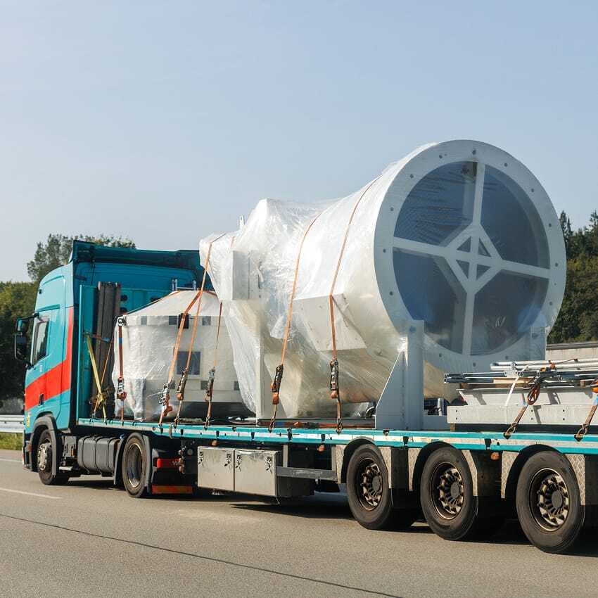 A large flatbed truck transports a wrapped cylindrical industrial component, secured with straps, along a highway bordered by trees under a clear sky.
