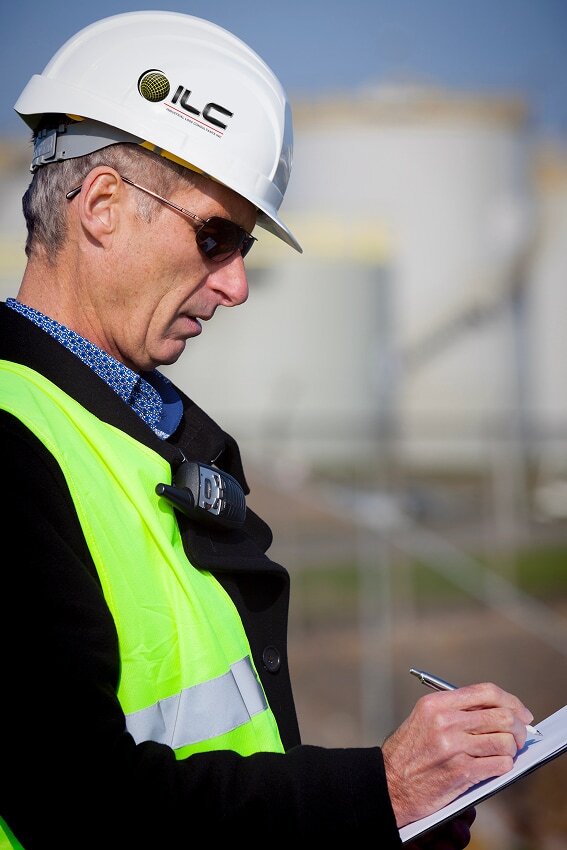 Man in a white hard hat and reflective vest, holding a clipboard, writes notes at a construction site. He appears focused and attentive.