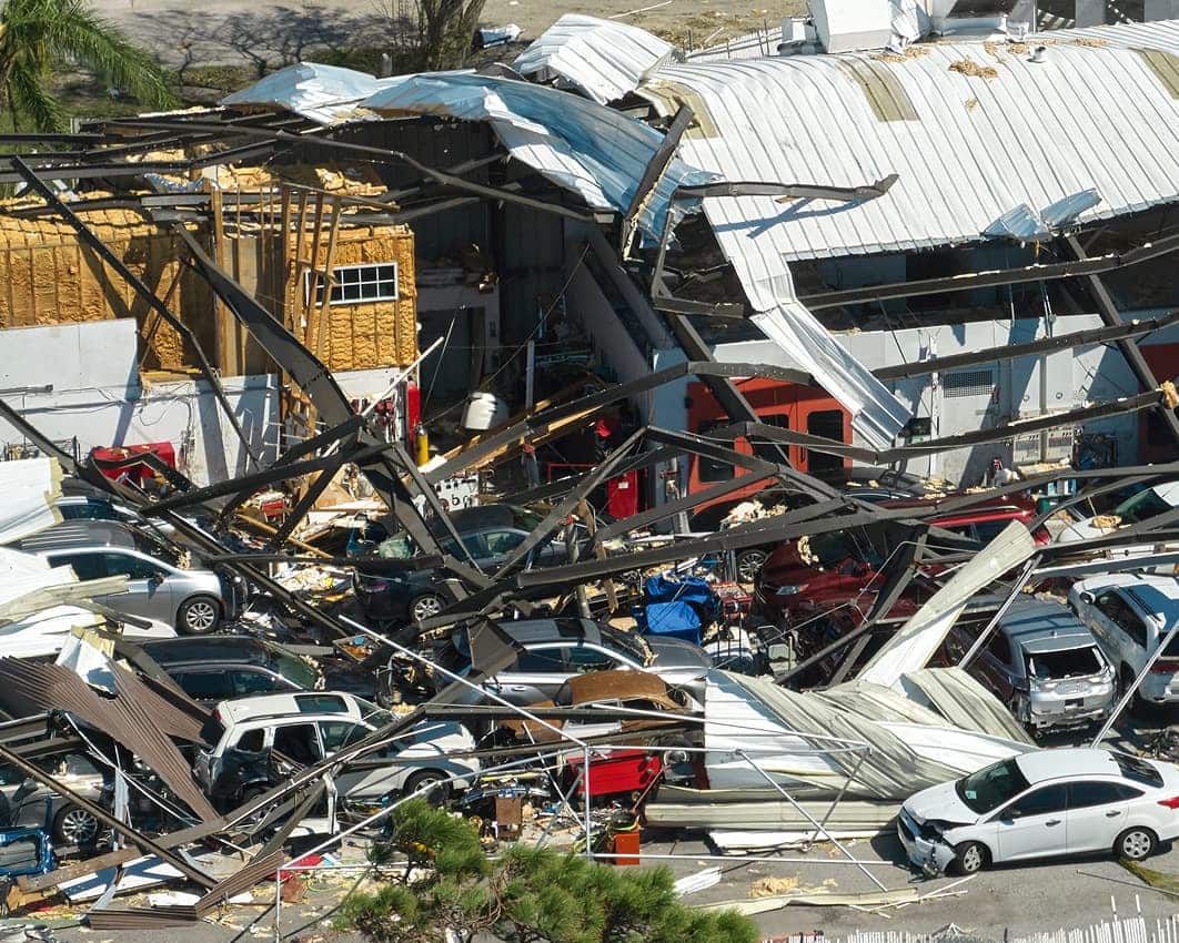 Collapsed metal roof and debris cover multiple cars in a parking lot. The scene suggests storm damage, depicting chaos and destruction.