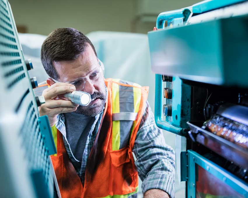 A worker in a safety vest and goggles inspects machinery with a flashlight. Focused expression, industrial environment, blue-green machine in foreground.