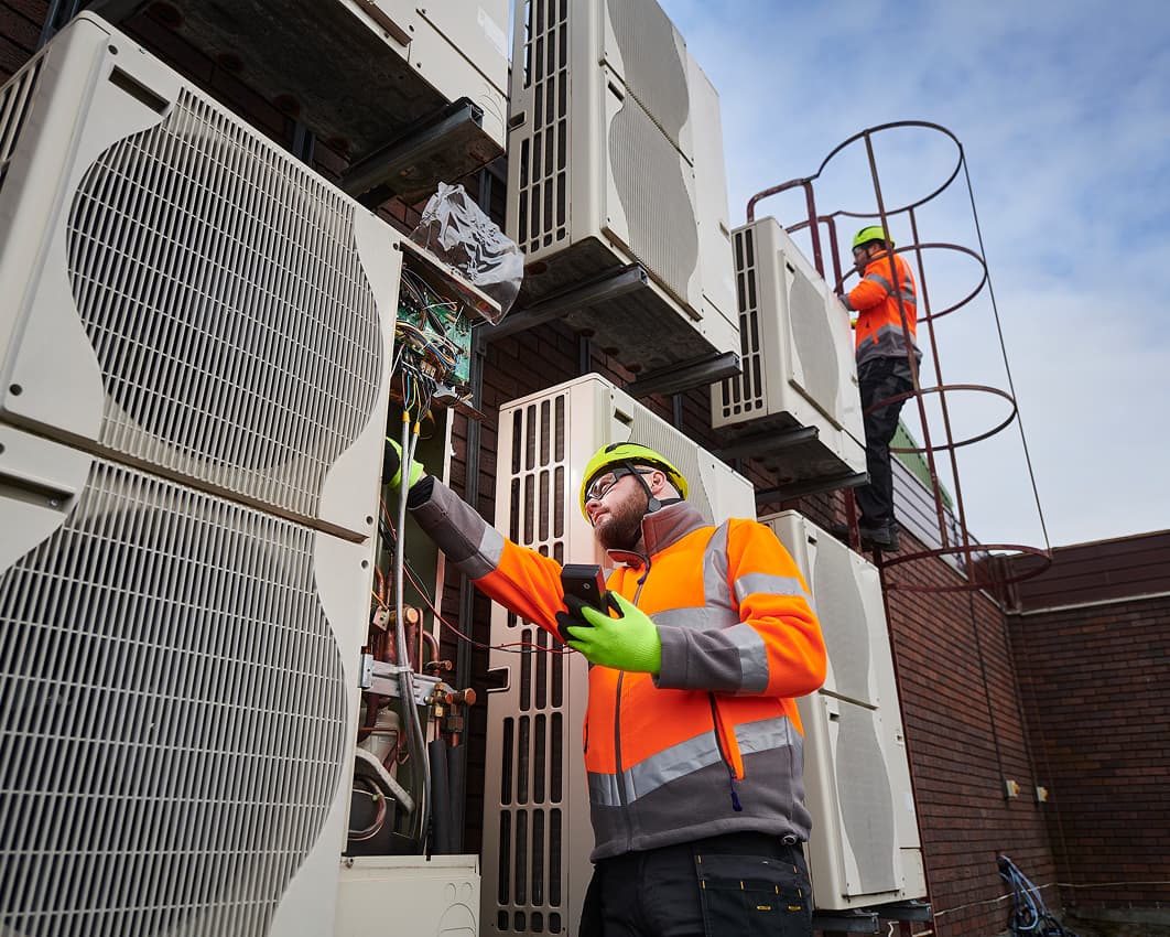 Two workers in high-visibility jackets and helmets inspect large outdoor air conditioning units mounted on a building; one is using tools, while the other stands on a metal ladder in the background.