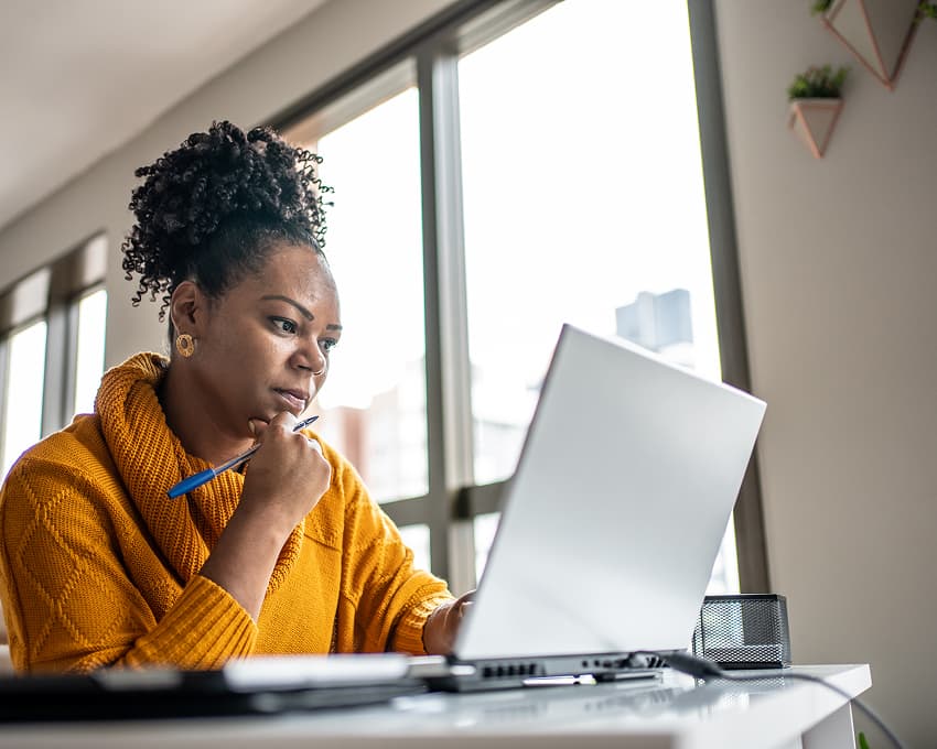 A woman in a cozy mustard sweater concentrates on a laptop in a bright, modern office. She's holding a pen, signifying thoughtfulness and focus.