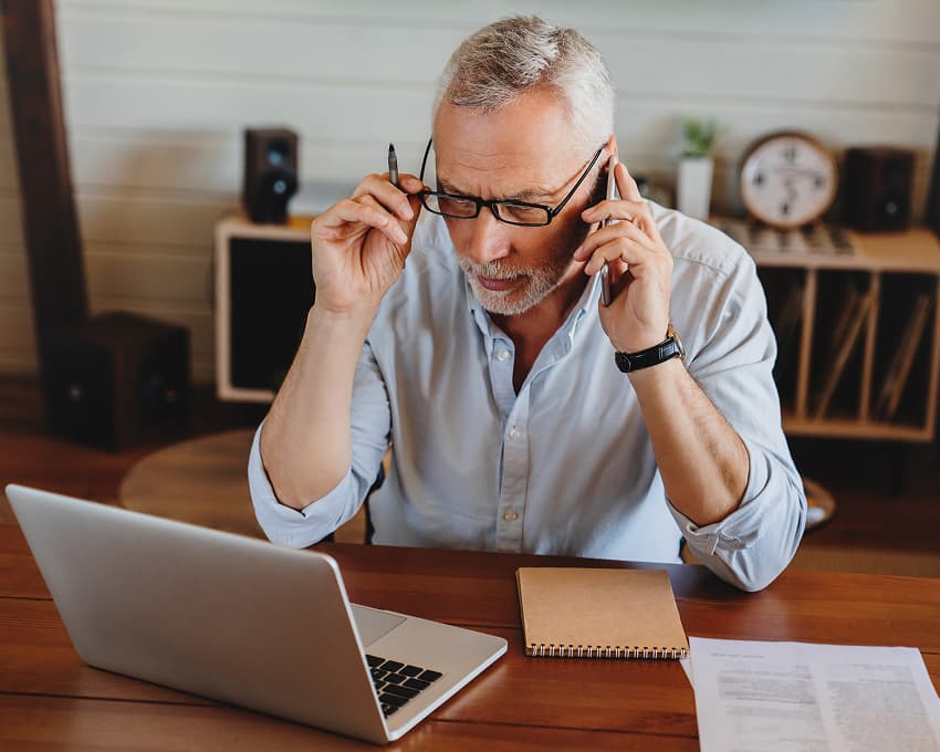 Elderly man with glasses at a wooden desk engaged in a phone call while looking at a laptop. He appears focused, surrounded by a notebook and papers.