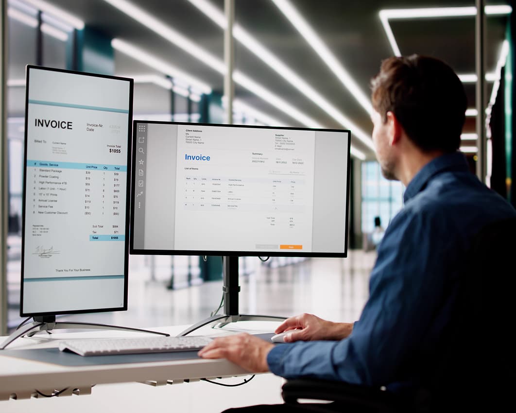A person in a blue shirt is working at a desk with two monitors displaying invoices, in a modern office with bright lighting and glass walls.