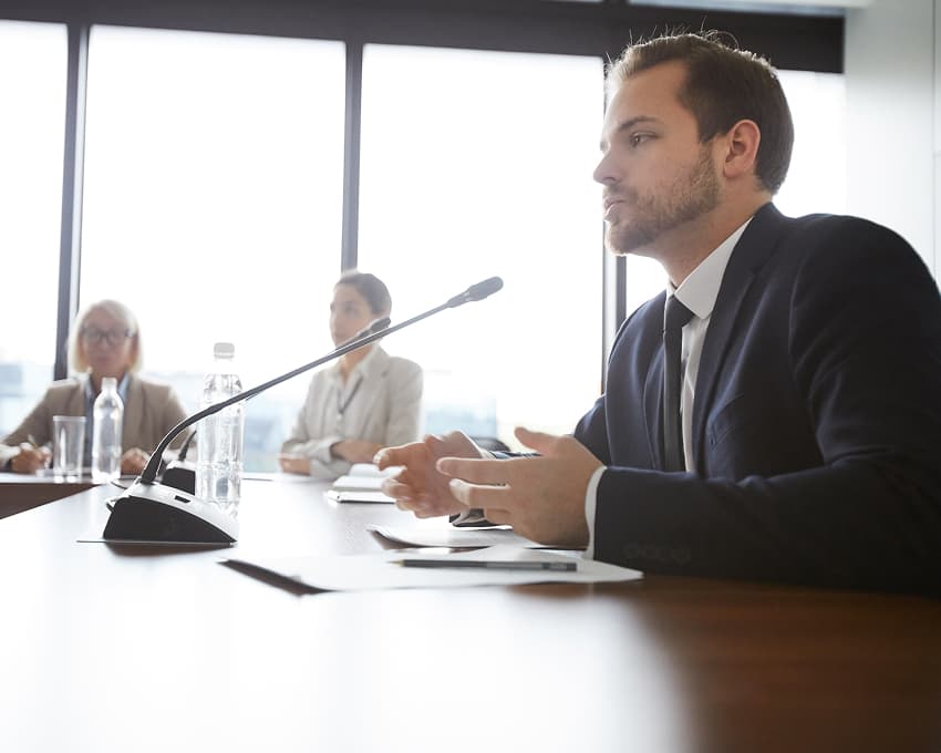 A man in a suit speaks into a microphone at a conference table, facing a group of attentive colleagues, set against bright windows. Professional atmosphere.