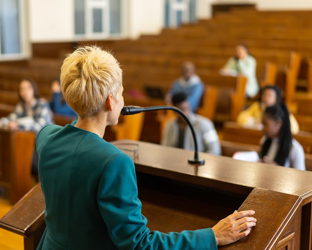 A person with short blond hair speaks at a podium in a lecture hall. Blurred students sit in the background, suggesting a focused, educational setting.