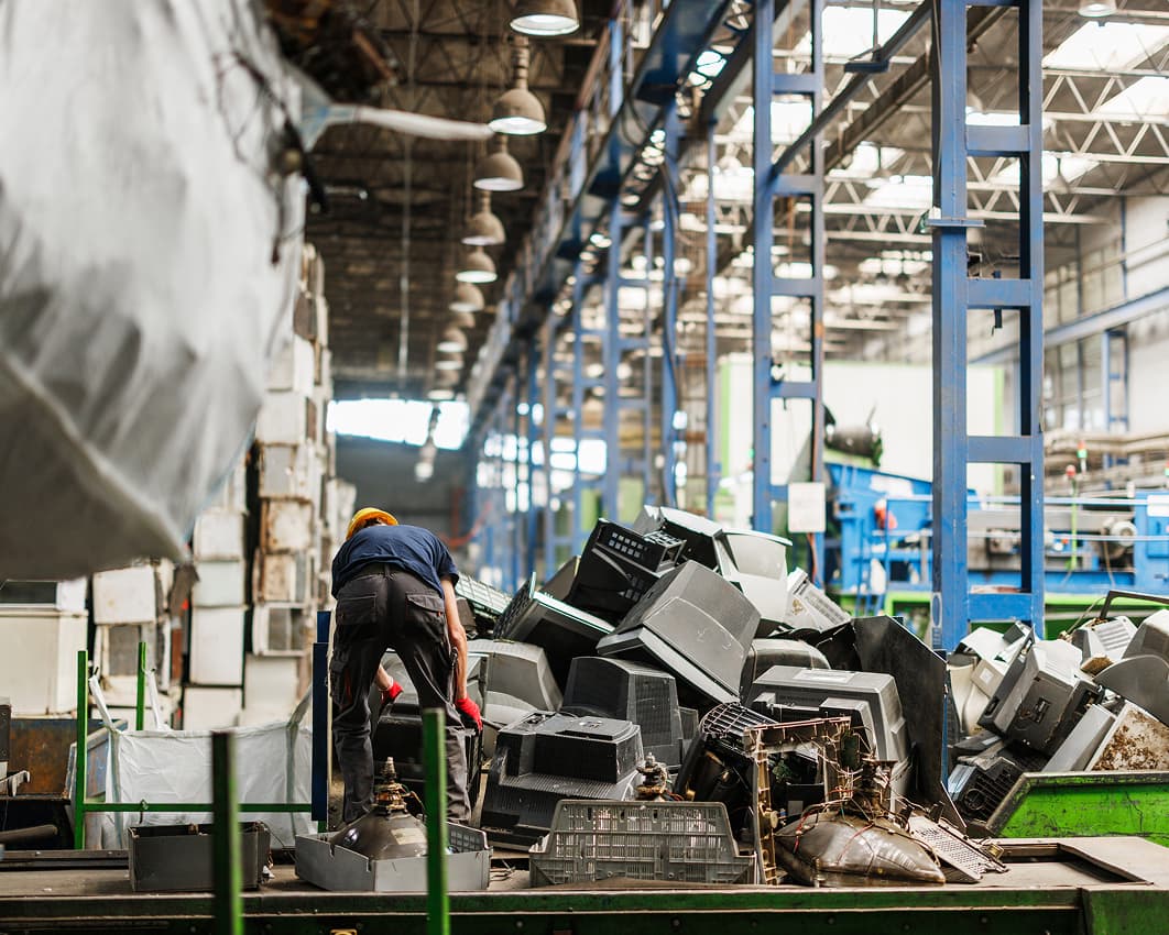 Worker in a large, industrial warehouse sorts through a heap of discarded electronics, including monitors and TVs, under bright overhead lights.