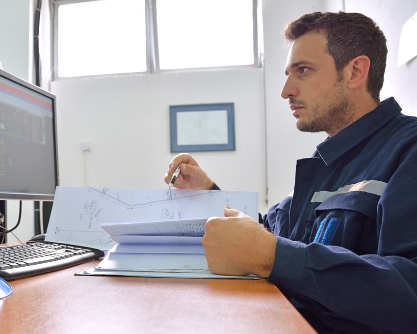 A man in a work uniform studies blueprints at a desk, using a computer with technical diagrams. A blue helmet sits nearby, conveying focus and professionalism.
