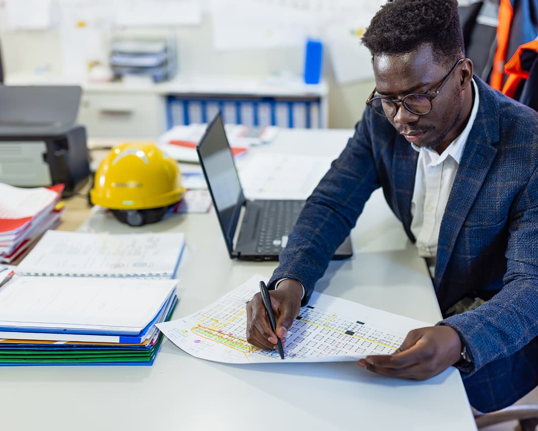 A man in a suit is working at a desk, reviewing technical documents. A laptop, hard hat, and safety vest are nearby, suggesting a construction setting.