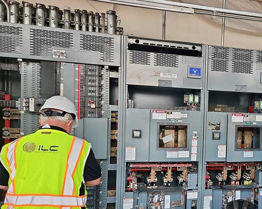An ILC consultant wearing a hard hat and high-visibility vest examines a large industrial electrical panel with open compartments, exposed wiring, switches, and breakers inside a mechanical or electrical room.
