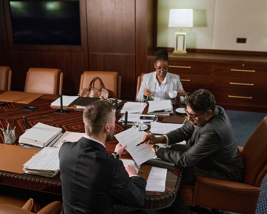 Business people collaborating in a meeting room, seated around a table with documents and laptops in front of them.
