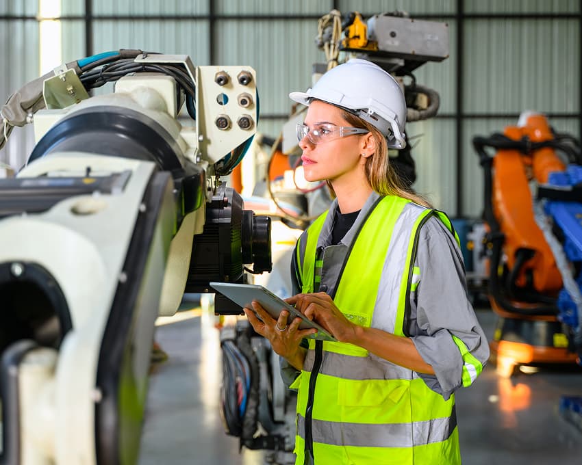A woman in a hard hat, safety glasses, and a high-visibility vest inspects industrial machinery in a factory, using a tablet. Robotic arms and equipment are visible in the background.