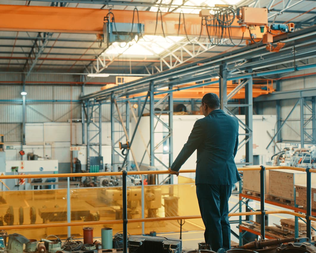 A man in a suit stands on an elevated walkway overlooking a large industrial factory floor with machinery, equipment, and overhead cranes.