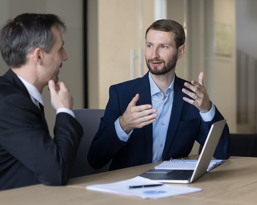 Two men in business attire sit at a table with a laptop and documents, engaged in a conversation in an office setting.
